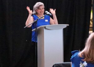 A woman in a blue dress at a podium raises her hands and smiles